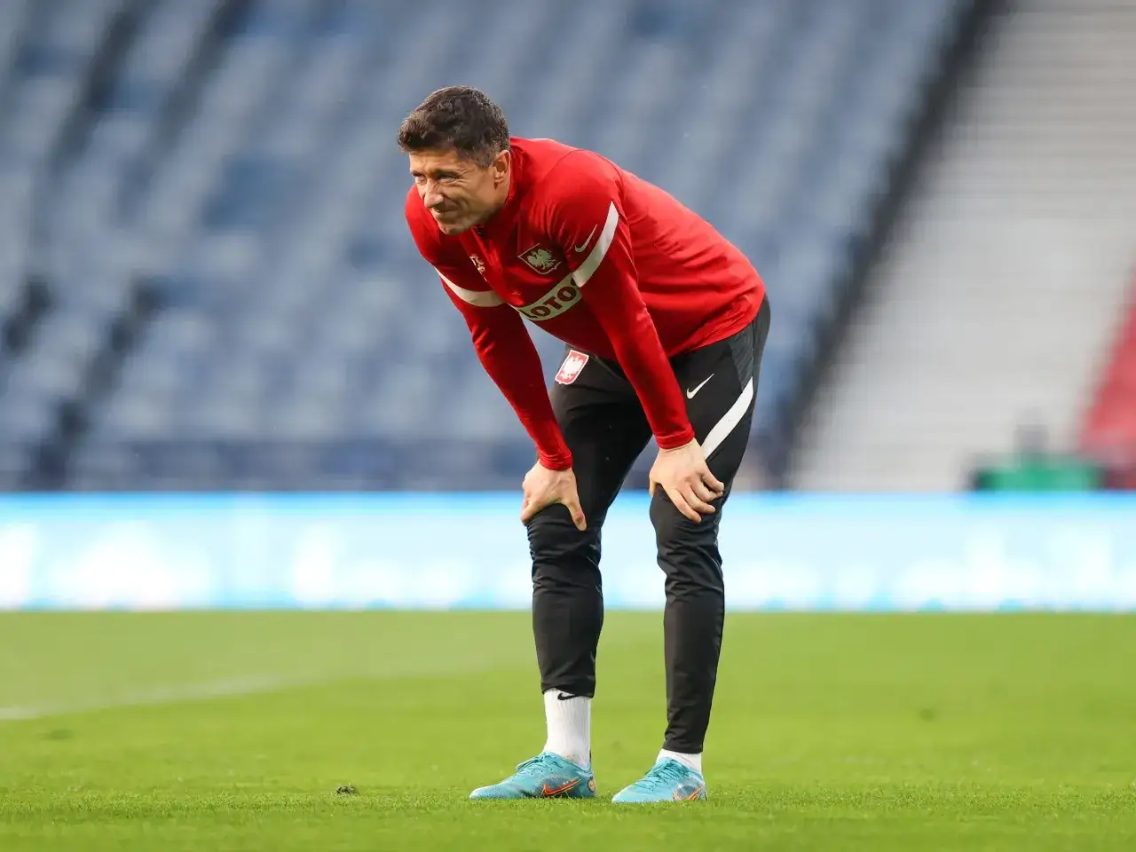 Piłkarz w czerwonej bluzie z orłem na piersi pochyla się, trzymając ręce na kolanach. Wygląda na to, że Robert Lewandowski odczuwa ból, być może to lewandowski kontuzja.