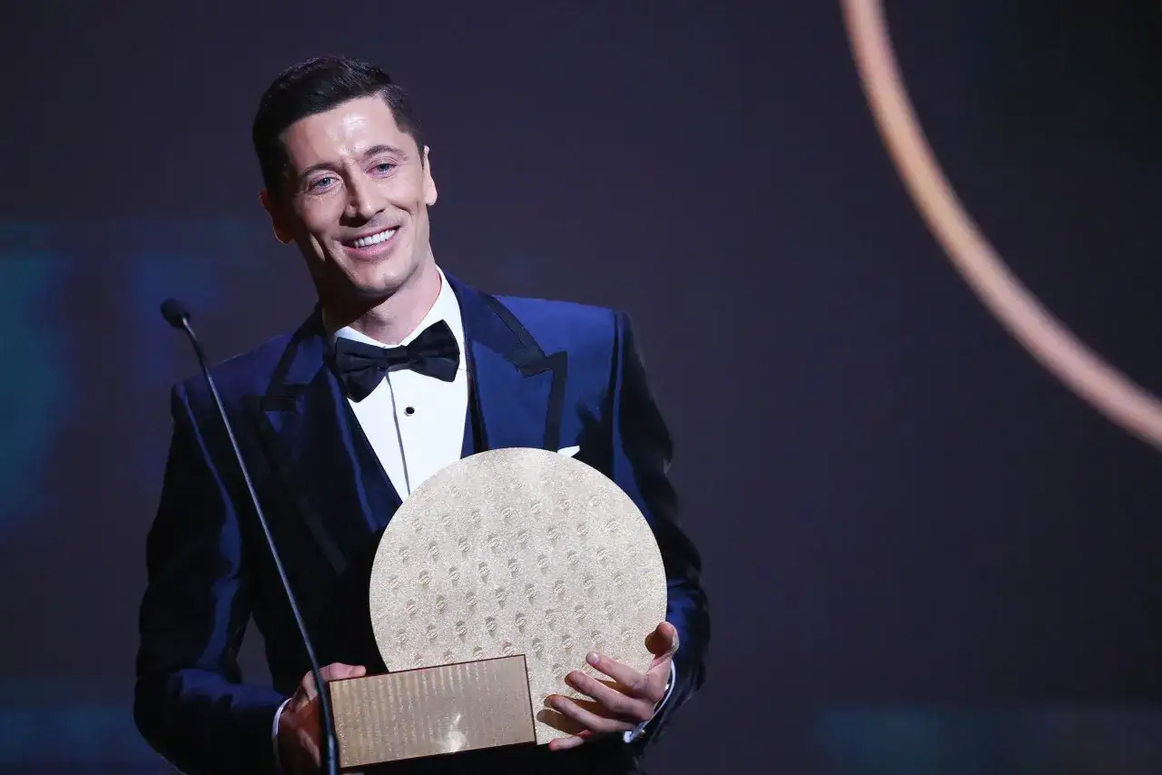 Uśmiechnięty Robert Lewandowski w eleganckim garniturze odbiera nagrodę, która przypomina Złotą Piłkę.