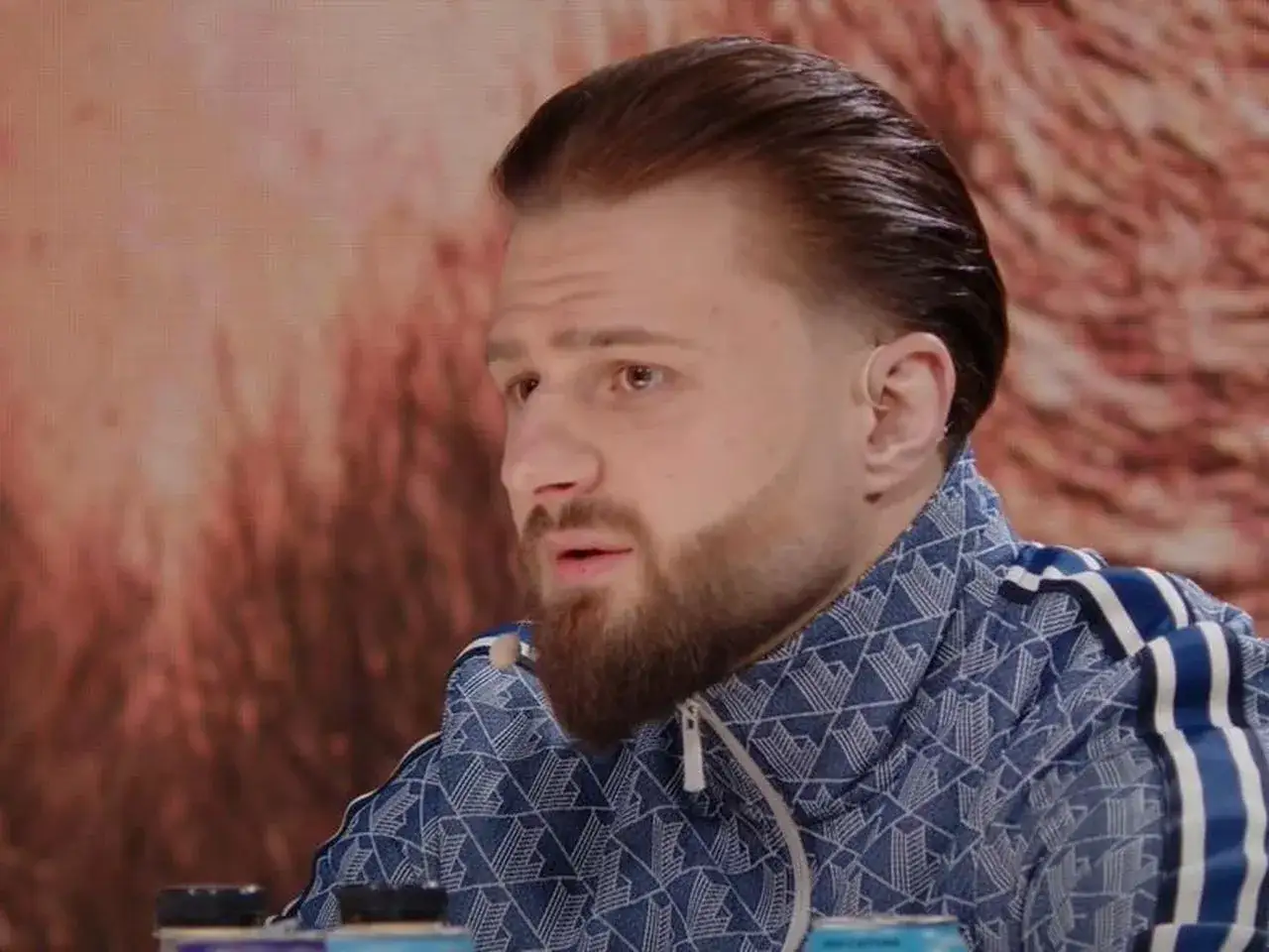 Amadeusz Ferrari w niebieskiej bluzie z białymi pasami, z charakterystyczną fryzurą i brodą, siedzi przed pomarańczowym tłem.