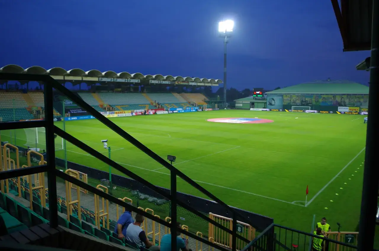 Wieczorny widok na stadion Górnik Łęczna. Piękne, zielone boisko i puste trybuny pod niebieskim niebem.