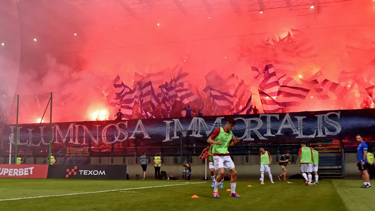 Piłkarze rozgrzewają się na boisku, podczas gdy kibice odpalają race i flagi na meczu forbet 4 liga warmińsko-mazurska.