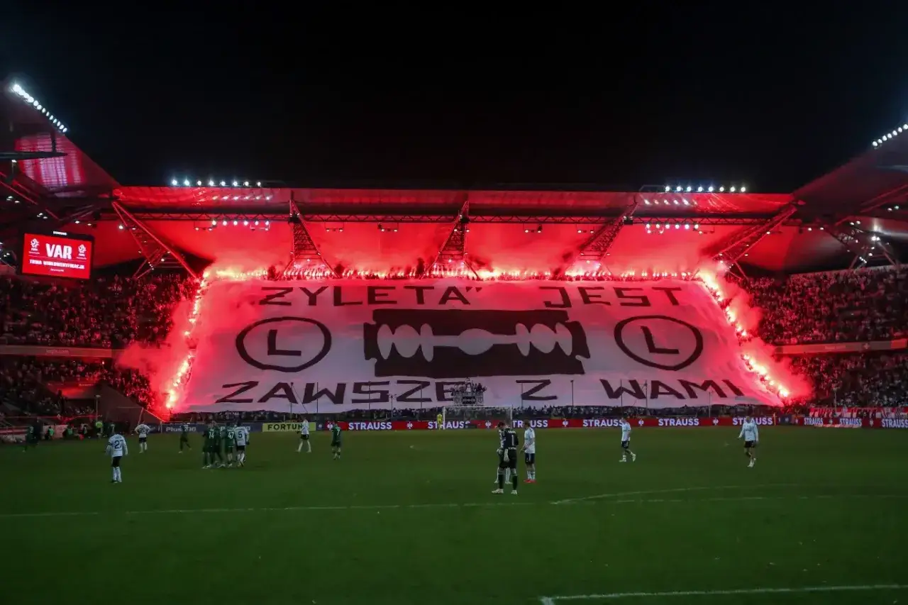 Trybuny stadionu Legii rozświetlone czerwonymi racami, ogromna oprawa z napisem 