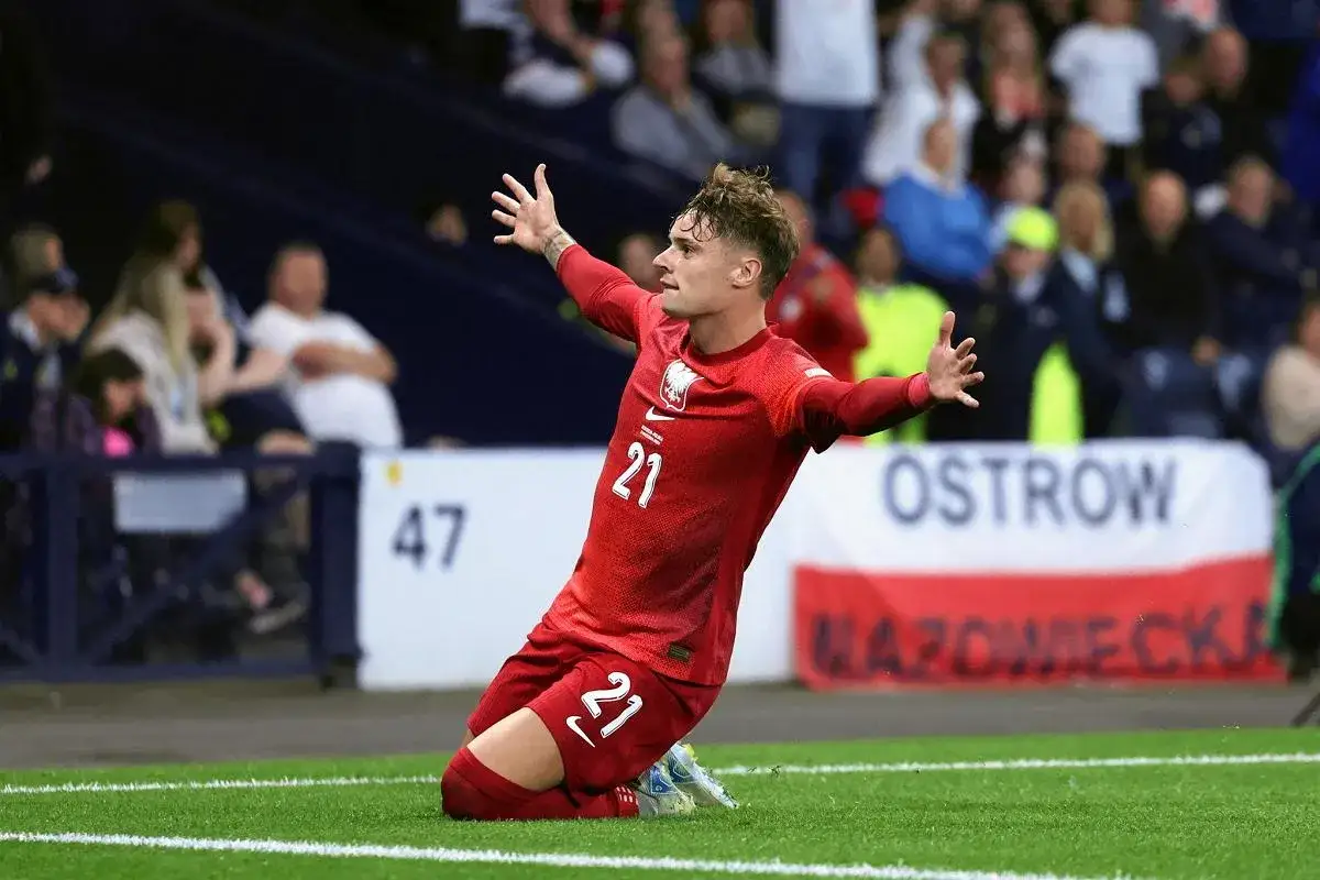 Piłkarz w czerwonej koszulce z numerem 21, Sebastian Szymański, klęczy na boisku z rękami uniesionymi w geście radości. Jego wzrost formy jest widoczny w tej dynamicznej pozie.