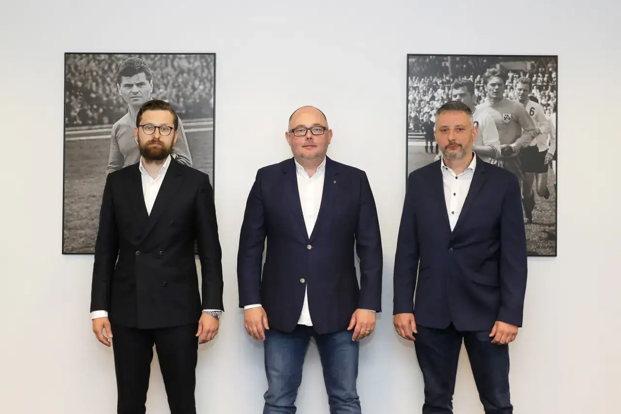 Trzech mężczyzn w garniturach przed czarno-białymi zdjęciami piłkarzy. Jeden z nich to prezes Legii.