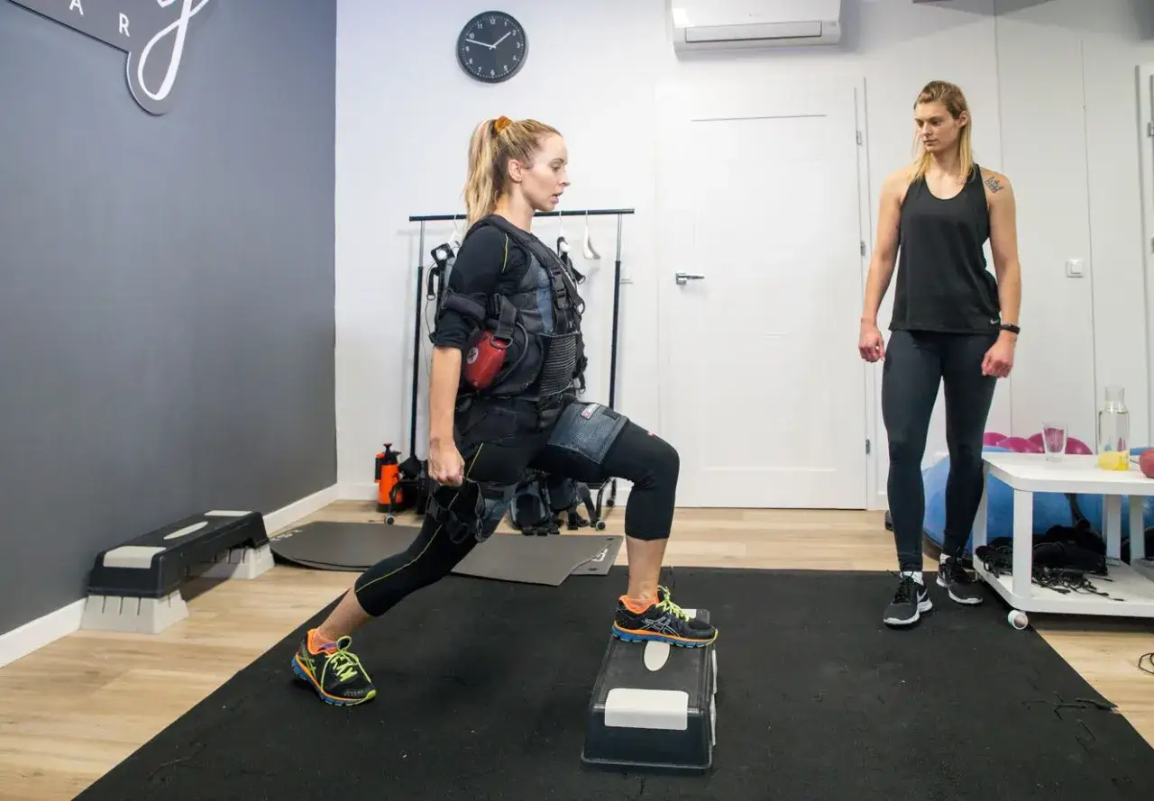 Kobieta w kamizelce EMS wykonuje wykrok, podczas gdy trenerka obserwuje. Zastanawiasz się, ile kosztuje trening EMS?
