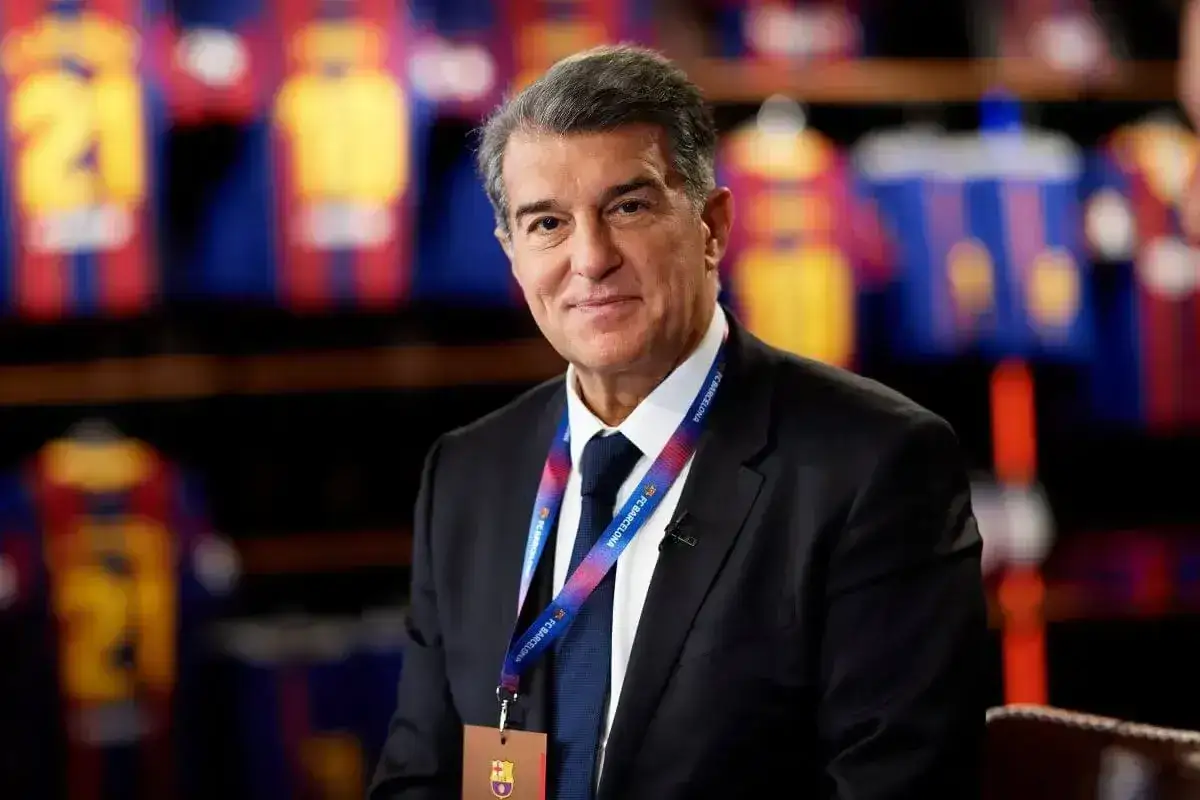 Uśmiechnięty prezes Barcelony, Joan Laporta, w garniturze, z identyfikatorem FC Barcelona na szyi. W tle widoczne koszulki piłkarskie.
