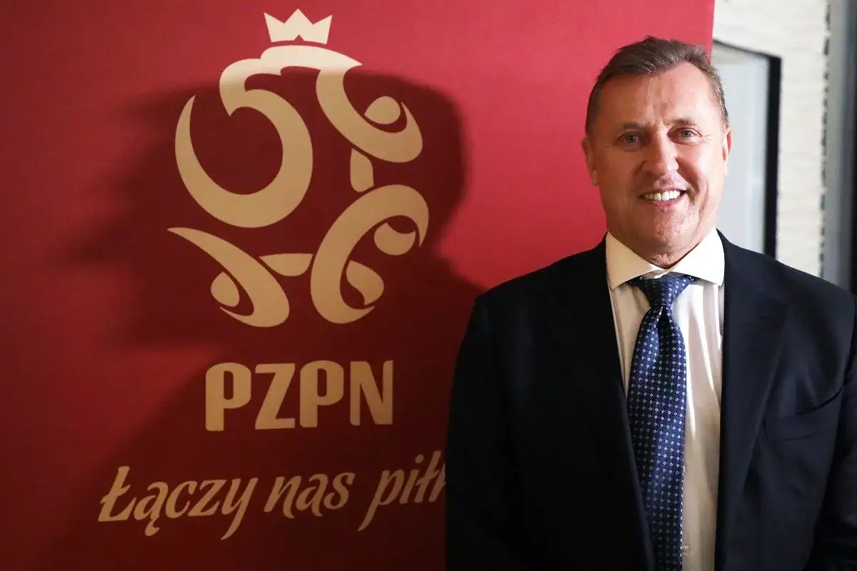 Uśmiechnięty prezes PZPN w garniturze stoi przed czerwonym tłem z logo związku.