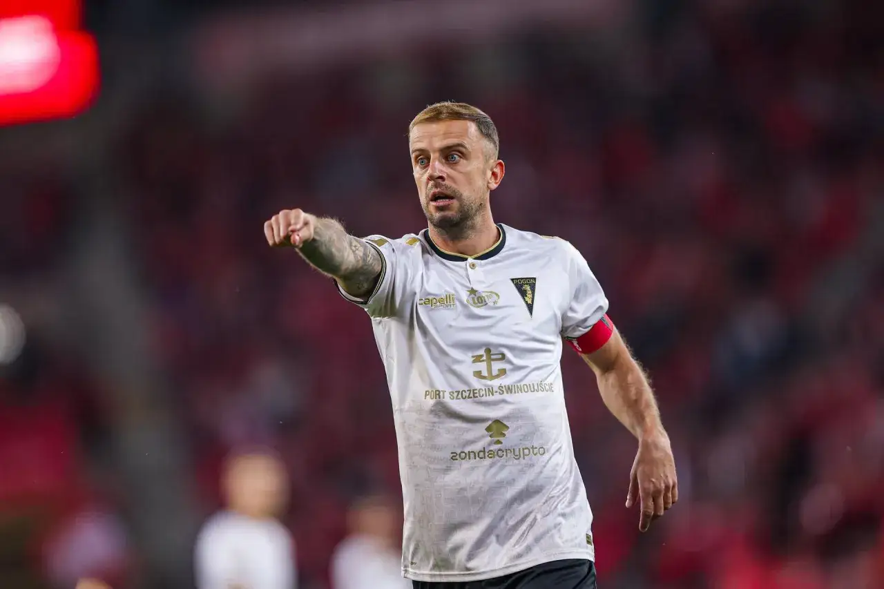 Kamil Grosicki w białej koszulce Pogoni Szczecin, z charakterystycznym tatuażem na ramieniu, wskazuje palcem. Jego obecne drużyny to Pogoń.