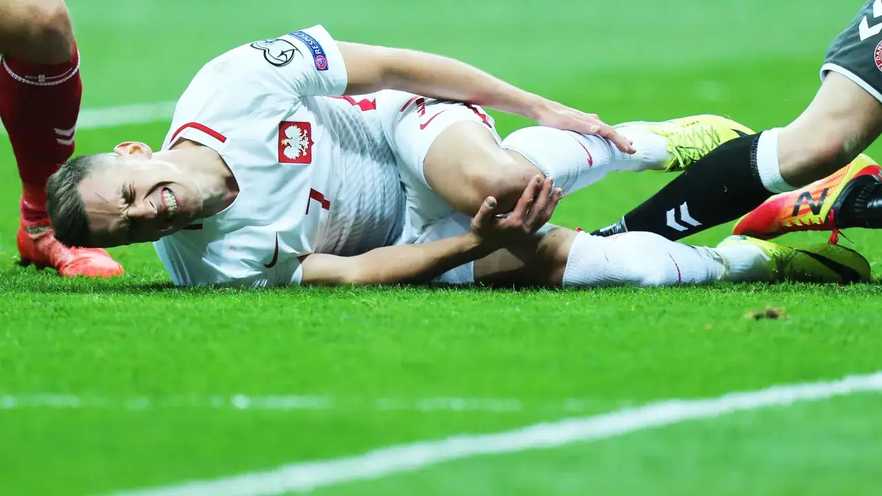 Piłkarz w białej koszulce z orłem na piersi leży na trawie, trzymając się za nogę. Widać jego grymas bólu, sugerujący poważną kontuzję.