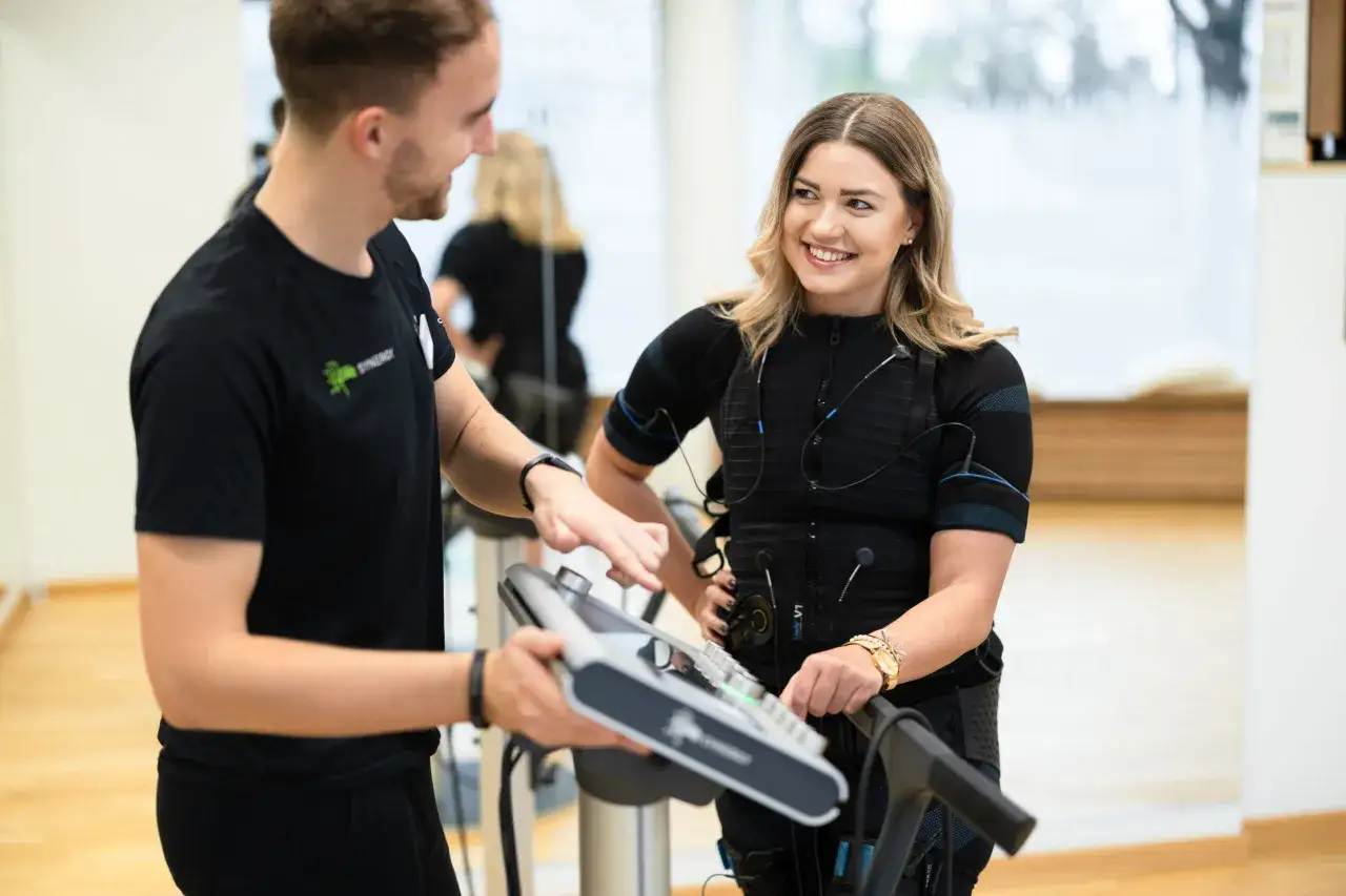 Trener pokazuje kobiecie w kamizelce EMS, ile kosztuje trening EMS. Uśmiechnięta para w centrum fitness.
