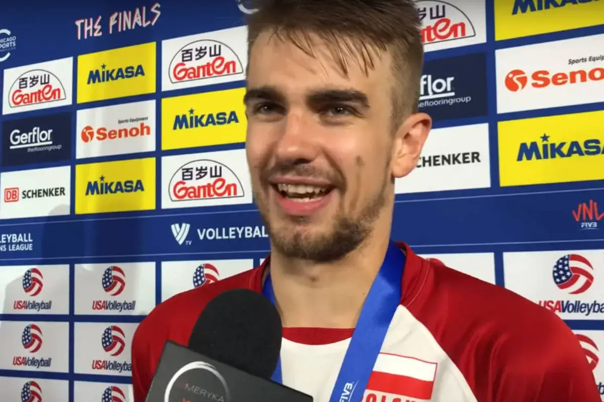Uśmiechnięty libero reprezentacji Polski po wygranym meczu. W tle logo sponsorów turnieju.