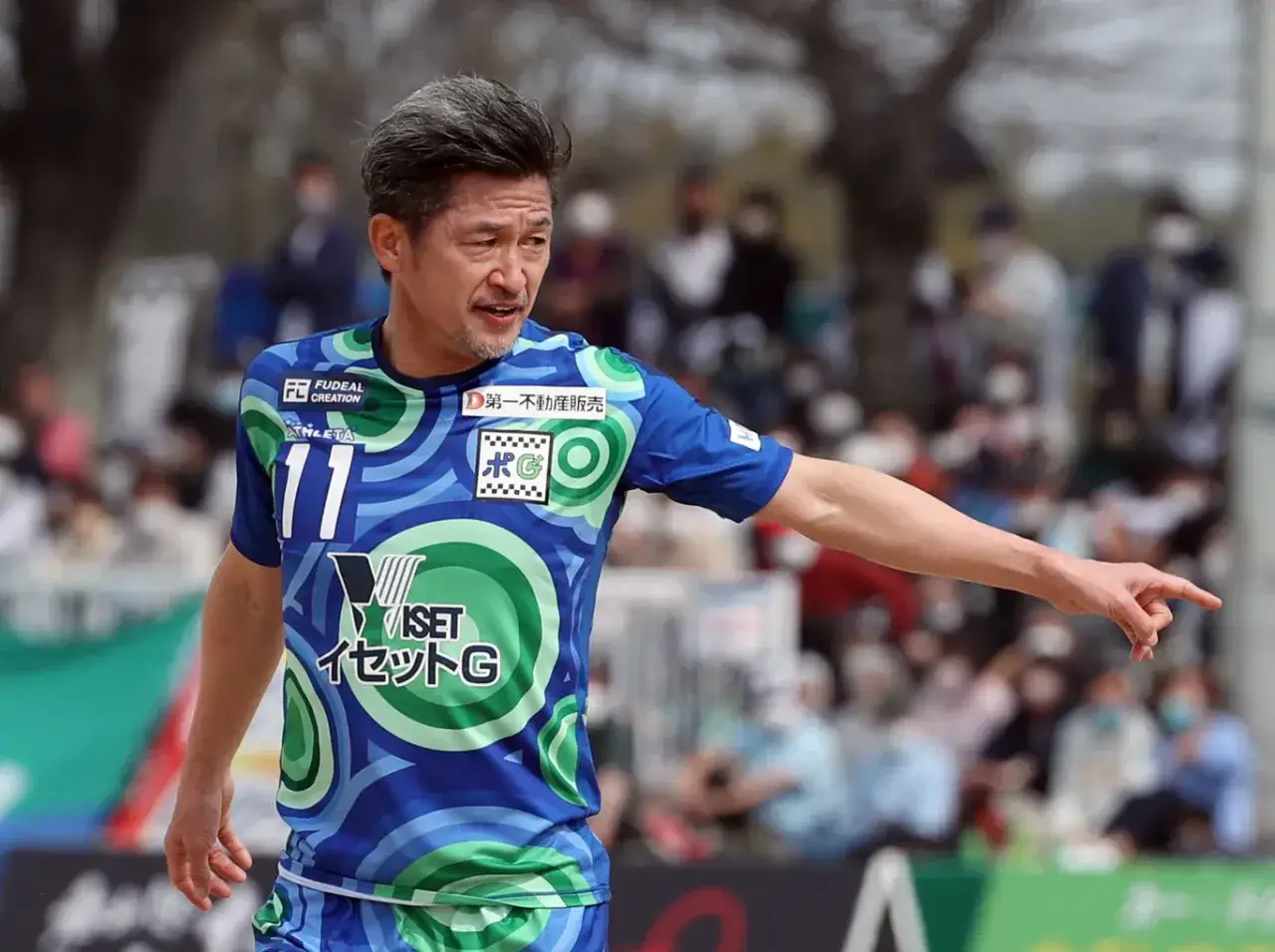 Najstarszy piłkarz na świecie, Kazuyoshi Miura, w niebieskiej koszulce z zielonymi wzorami, wskazuje palcem.