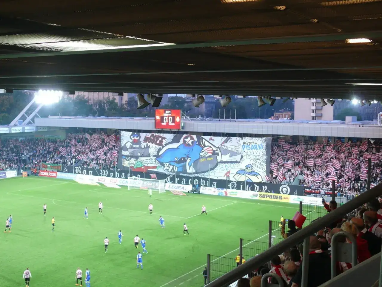 Widok stadionu podczas meczu piłki nożnej, gdzie kibice tworzą oprawę. To pokazuje, co to są derby w piłce nożnej – emocje i rywalizacja.