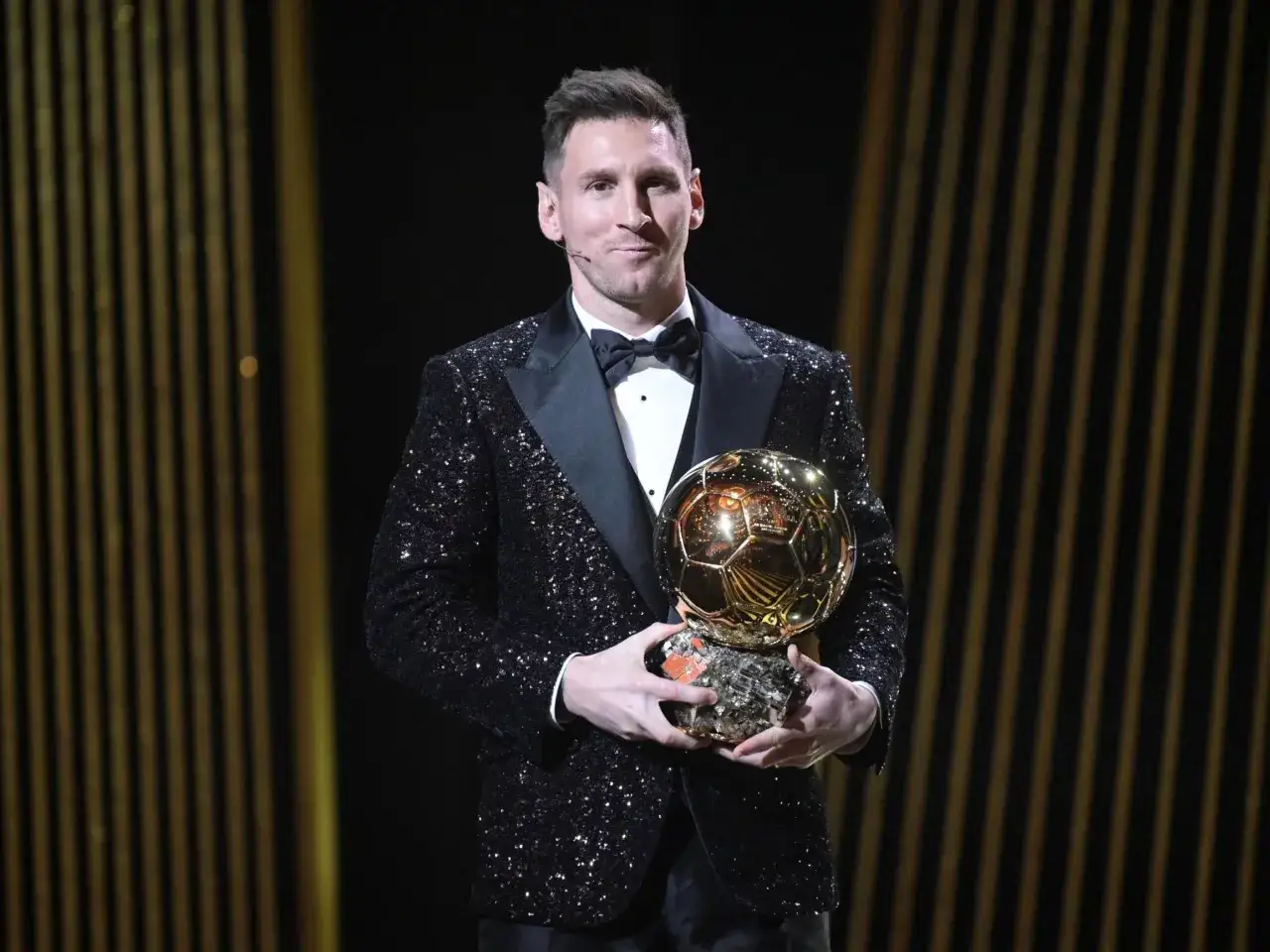 Lionel Messi trzyma Złotą Piłkę w błyszczącej marynarce na tle złotych pasów.