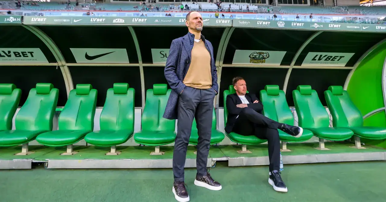 Trener Śląska Wrocław Ivan Djurdjevic stoi na tle zielonych siedzeń rezerwowych. Obok siedzi inny członek sztabu.