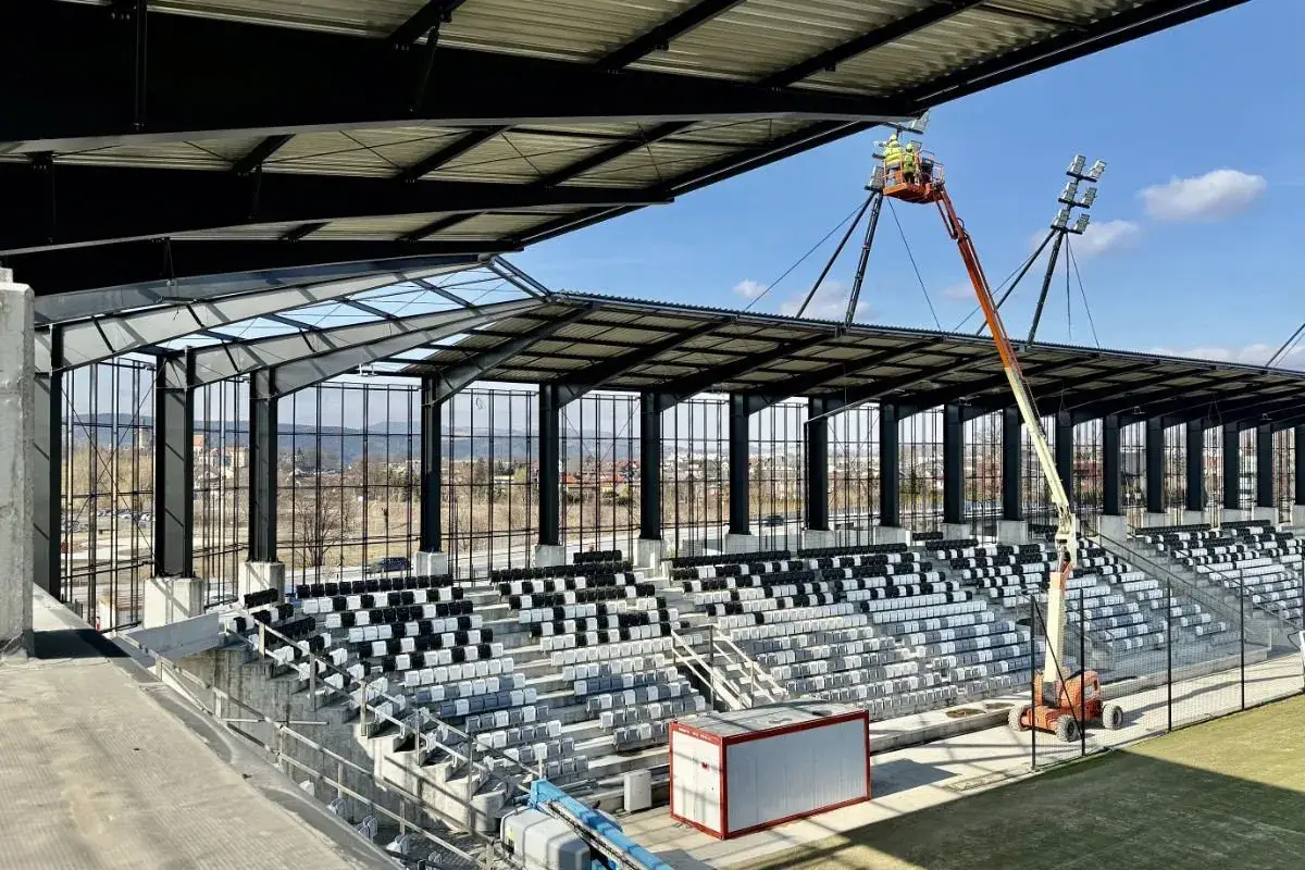 Trwa modernizacja stadionu Sandecji. Widać nowe trybuny, zadaszenie i maszty oświetleniowe. Pracownicy na podnośniku montują oświetlenie.