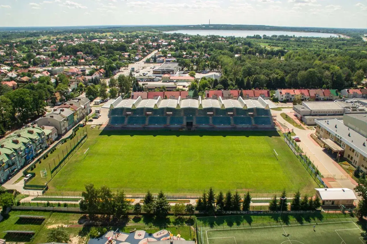 Stadion Siarka Tarnobrzeg z zieloną murawą i trybunami, otoczony zabudowaniami i lasem.