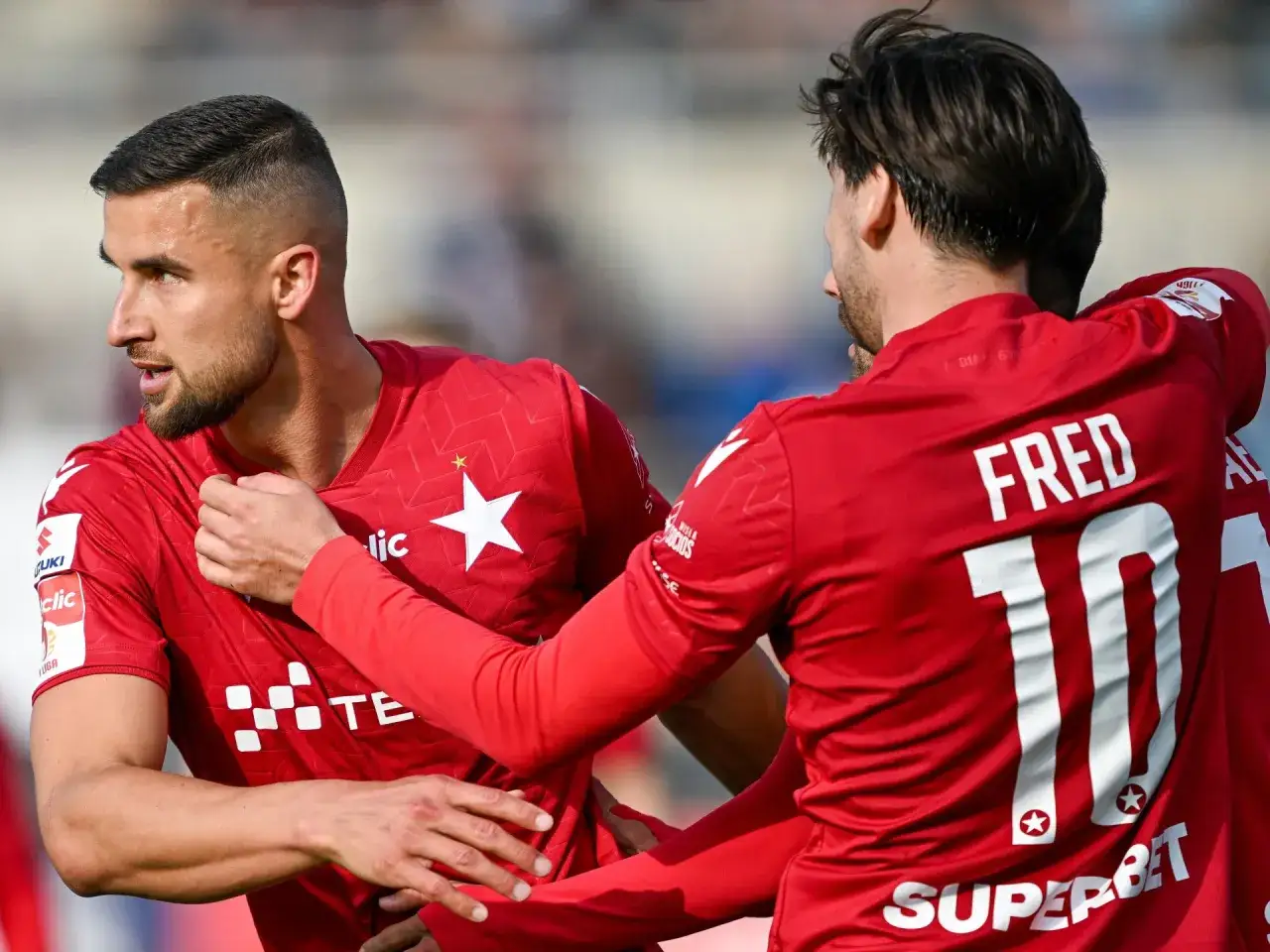 Piłkarze w czerwonych koszulkach świętują, być może awans do ekstraklasy. Na plecach jednego z nich widnieje napis