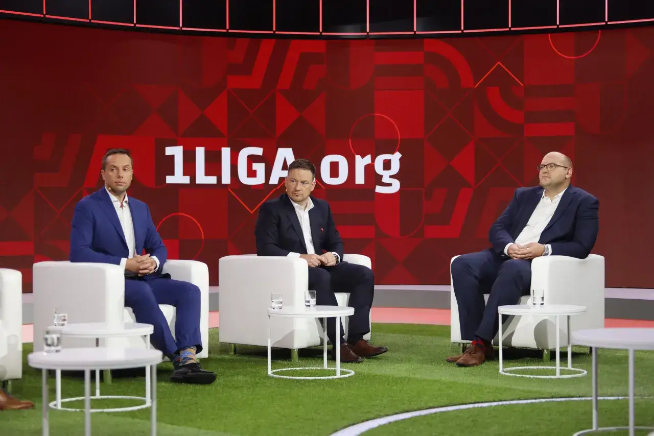 Trzech mężczyzn w garniturach siedzi na białych fotelach na tle napisu 