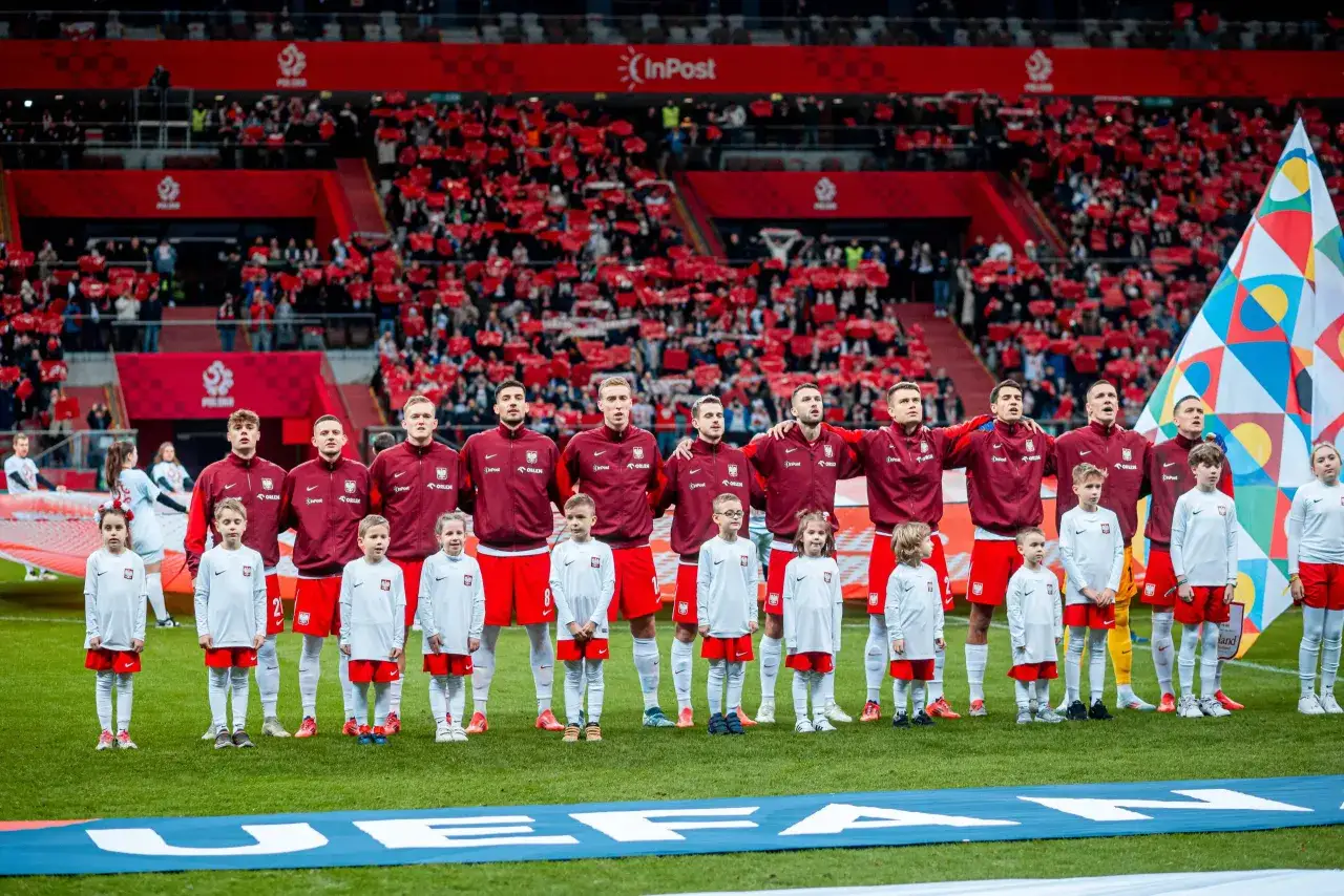 Piłkarze reprezentacji Polski i dzieci stoją na murawie stadionu, gotowi do meczu. Tłumy kibiców na trybunach czekają, kiedy gra polska w piłkę nożną.