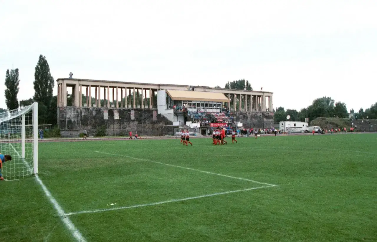 Mecz na starym stadionie Wisły Kraków. Piłkarze w czerwonych strojach rozgrywają spotkanie na zielonej murawie.