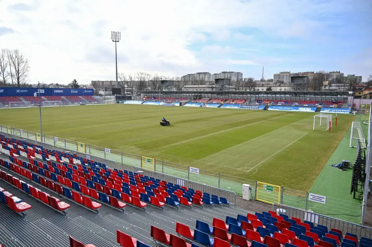 Pusta trybuna stadionu z czerwonymi i niebieskimi siedzeniami. Na boisku widać traktorek do koszenia trawy. Czekamy na mecz Raków dzisiaj!