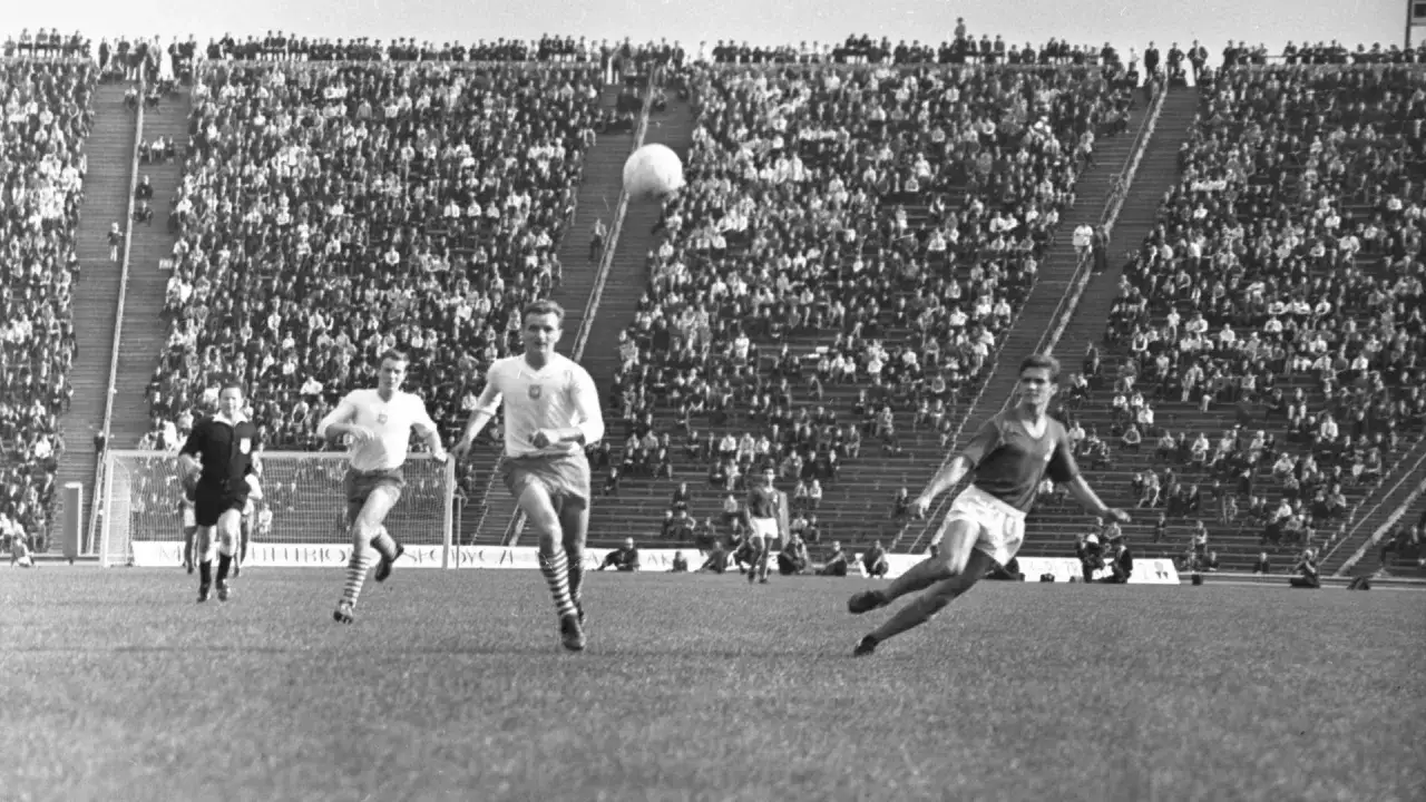 Mecz piłki nożnej na stadionie pełnym widzów. Piłkarze w akcji, jeden z nich biegnie z piłką. Pytanie 