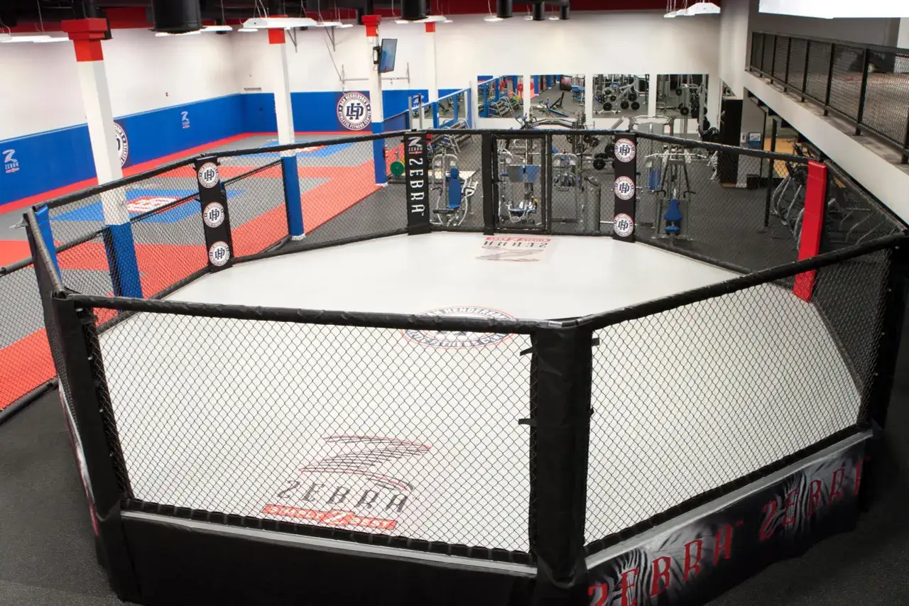 Pusta klatka MMA, gotowa na walkę. W tle siłownia i maty treningowe, czekające na zawodników MMA.