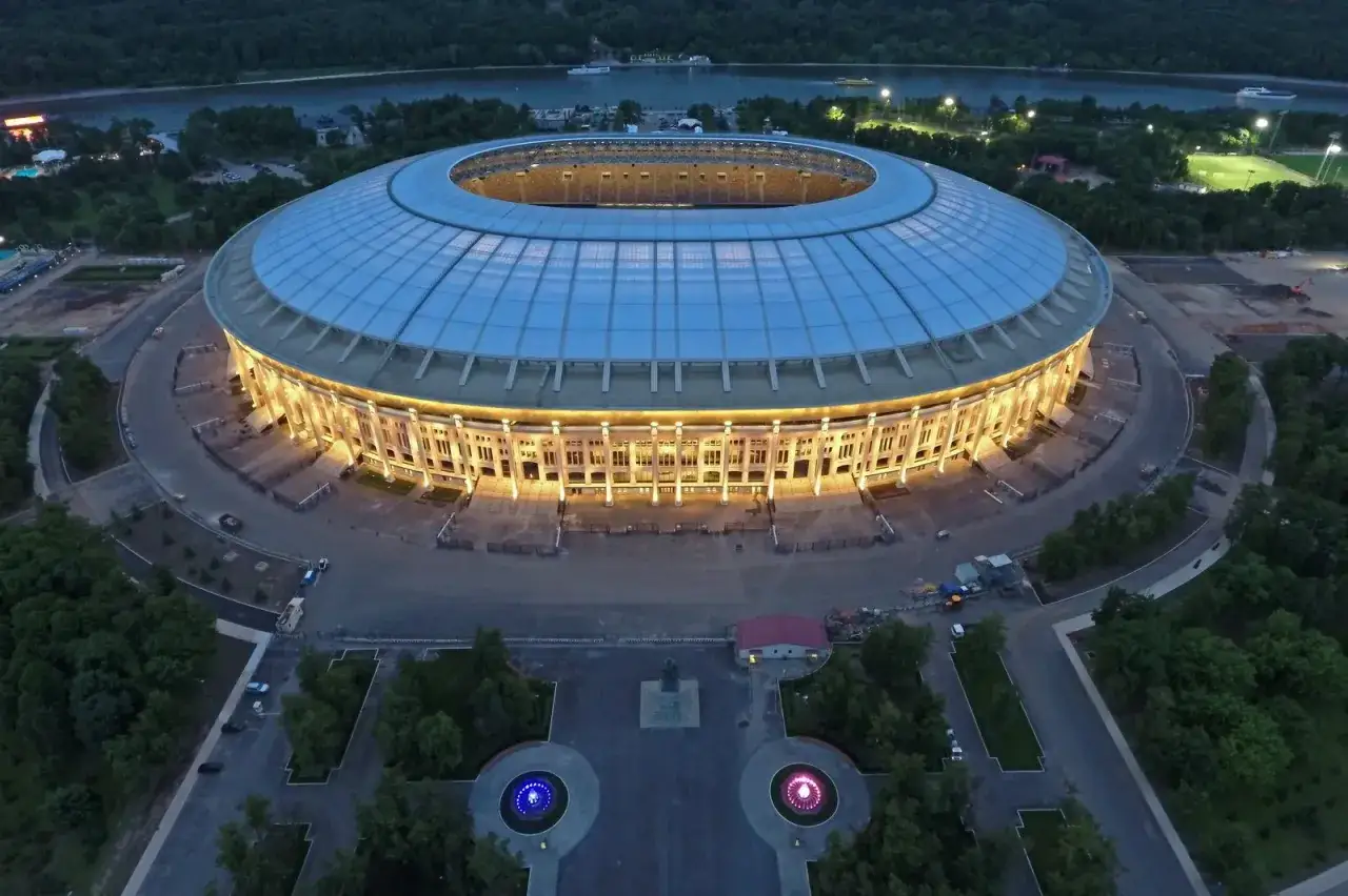 Oświetlony moskiewski stadion w nocy, z widokiem na rzekę i otaczający las.